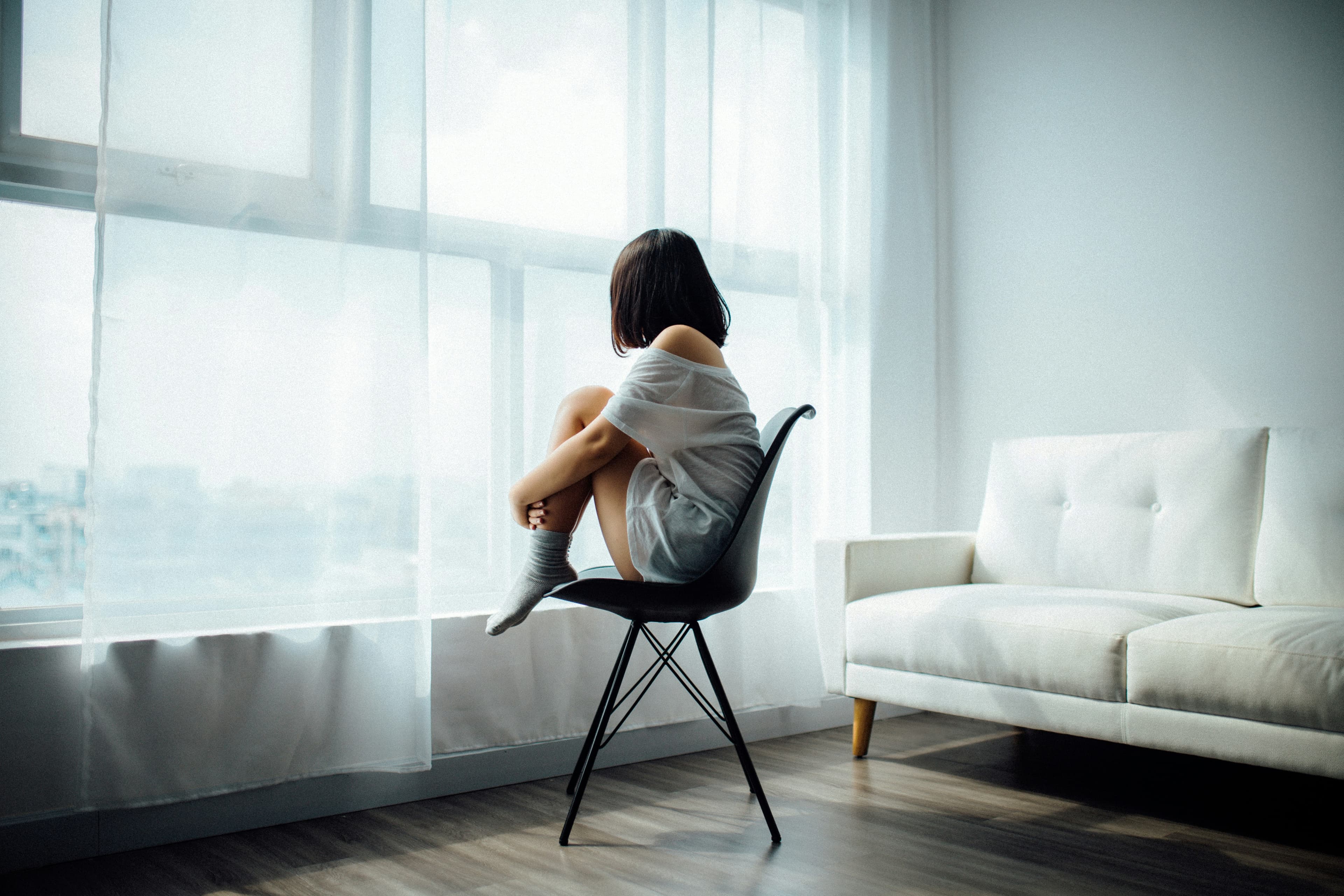 Mujer sentada sola frente a una ventana, abrazando sus rodillas
